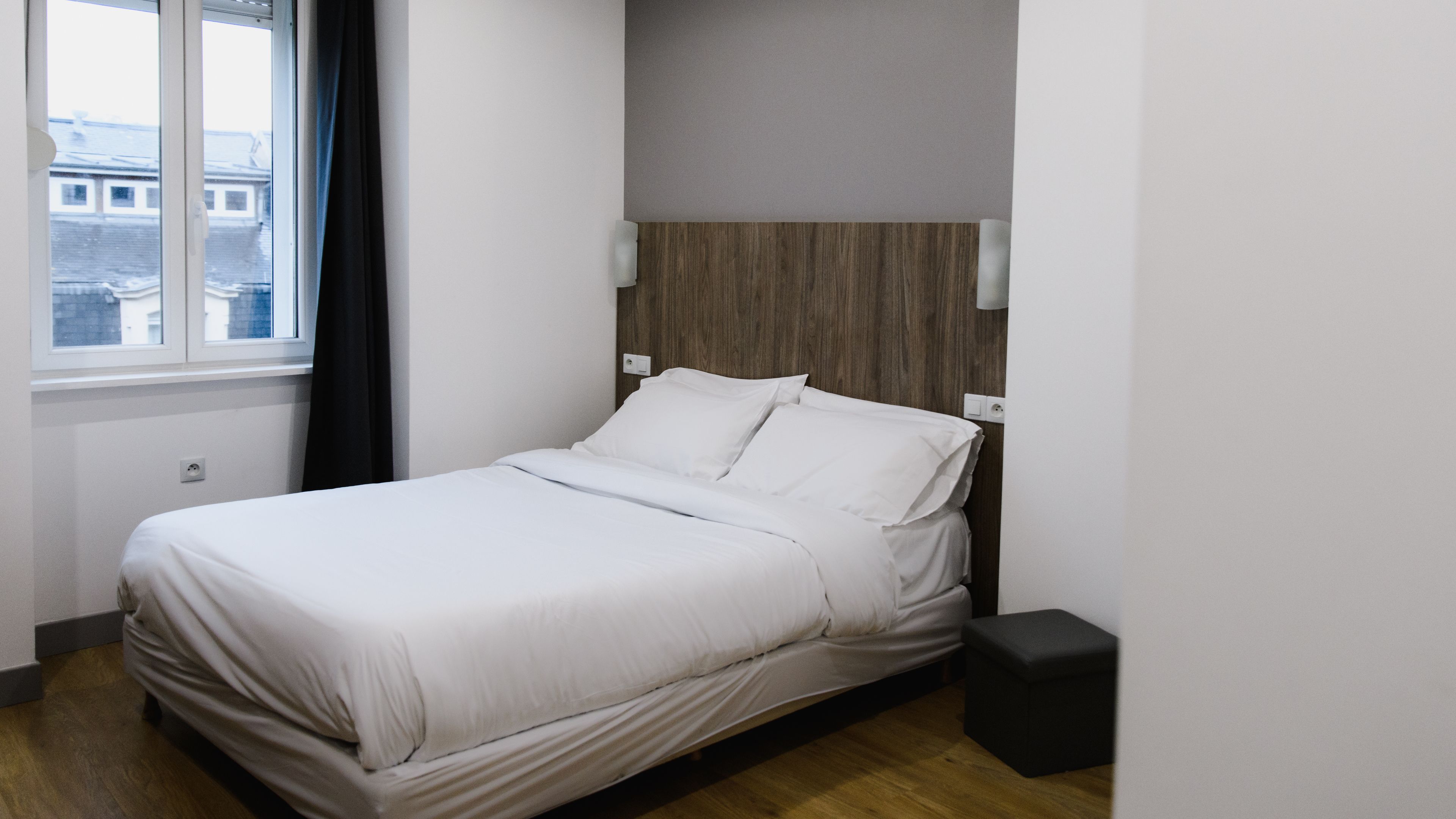 A neatly made bed in a room with a window, a wooden headboard, and a small bedside table.