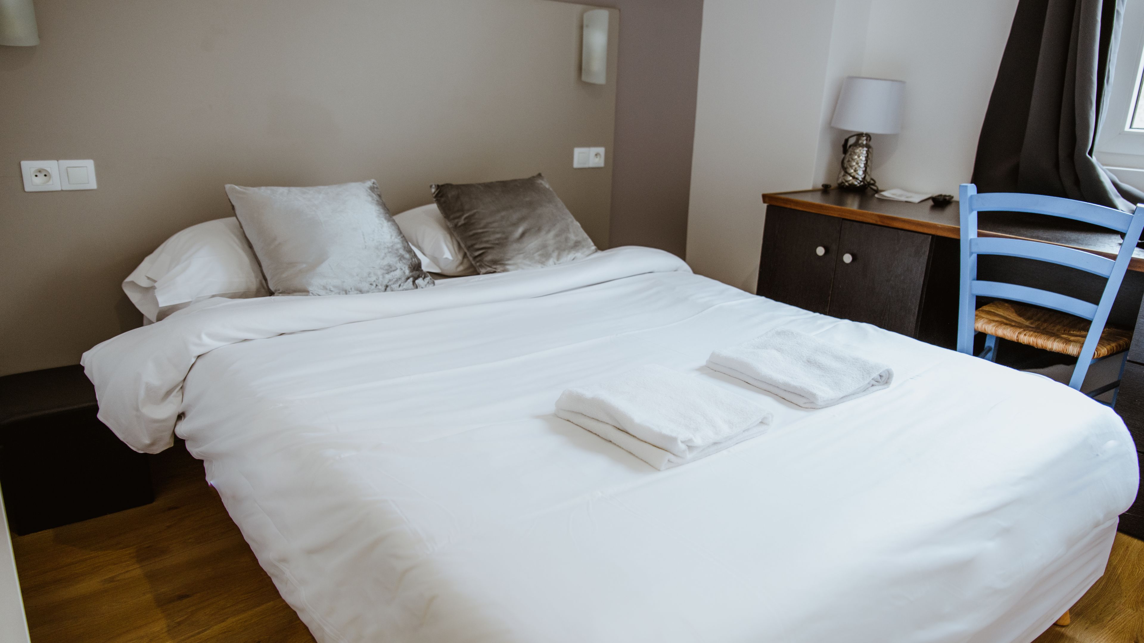 A neatly made bed with white linens and two pillows, a wooden nightstand with a lamp, and a blue chair.