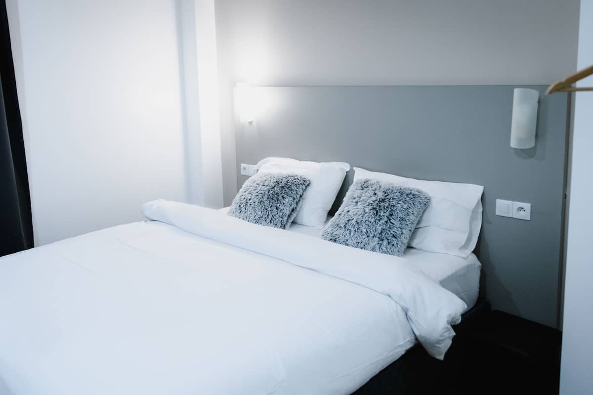 A neatly made bed with white linens and two fluffy pillows, a bedside table with a lamp, and a dark curtain on the left.