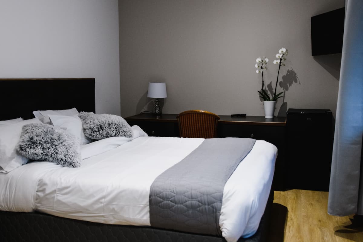A neatly made bed with a dark headboard, a bedside table with a lamp, a chair, and a desk with a plant.