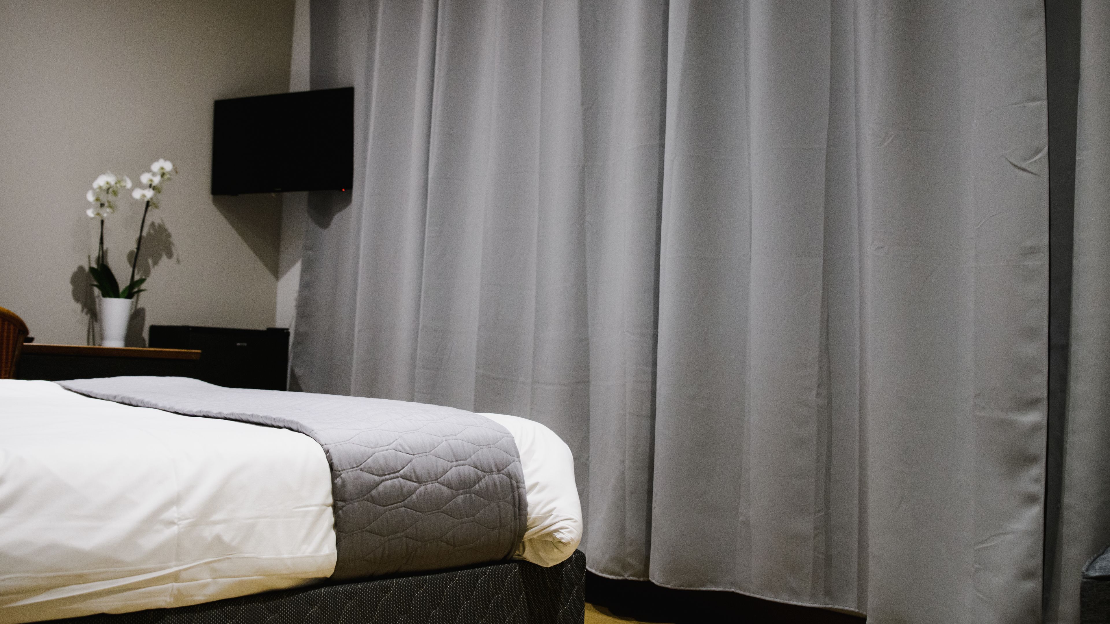 A neatly made bed with a quilted gray and white duvet, a small bedside table with a white vase and a potted plant, and a wall-mounted TV.