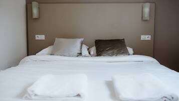 Una cama bien hecha con sábanas blancas y dos almohadas, una blanca y otra marrón oscuro, en una habitación con una pared lisa y dos lámparas fijadas a la pared.