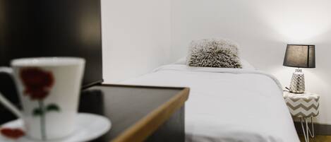 Un lit bien fait, avec un oreiller moelleux, une table de chevet sur laquelle se trouve une lampe, et une tasse ornée d’une rose.