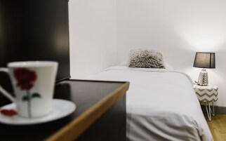 Una cama bien hecha con un almohadón mullido, una mesita de noche con una lámpara y una taza con una rosa.