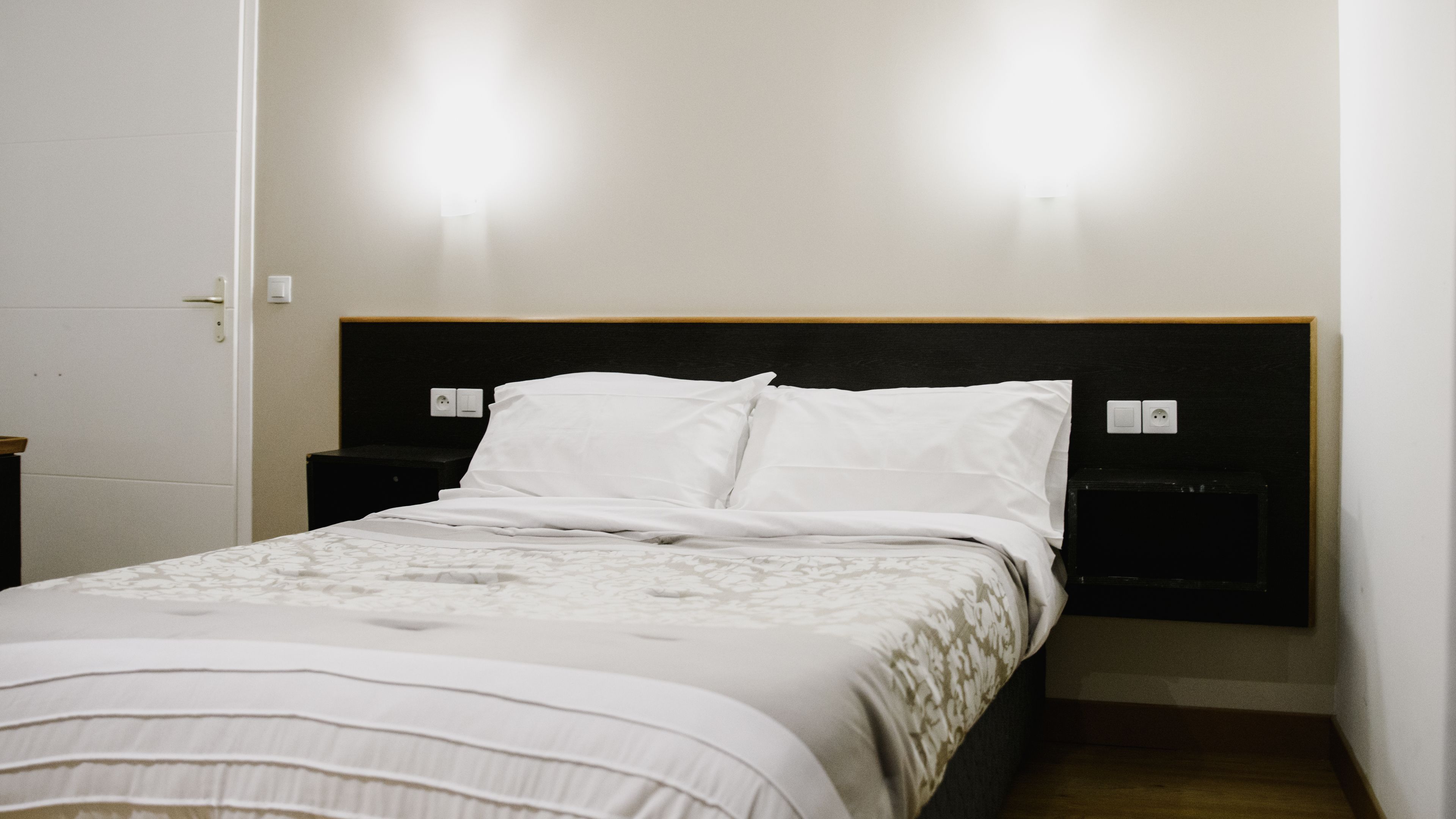 Un lit bien fait, avec du linge de lit blanc et une couette à motifs, une tête de lit noire et deux tables de chevet équipées de prises électriques murales.