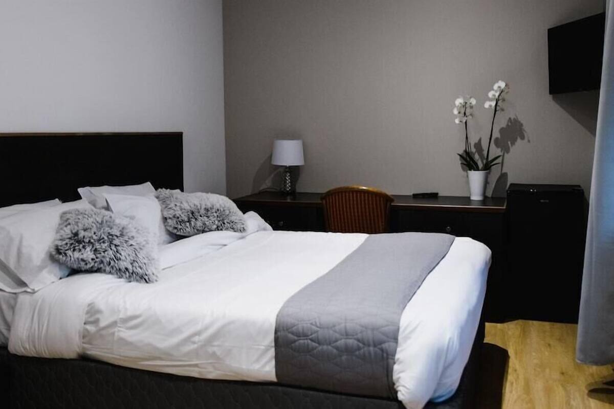 A neatly made bed with white and gray bedding, a dark headboard, a small desk with a lamp, a chair, and a potted plant.