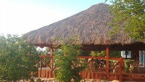 Una estructura con techo de paja y barandillas de madera, rodeada de árboles y arbustos pequeños, con una montaña como fondo.