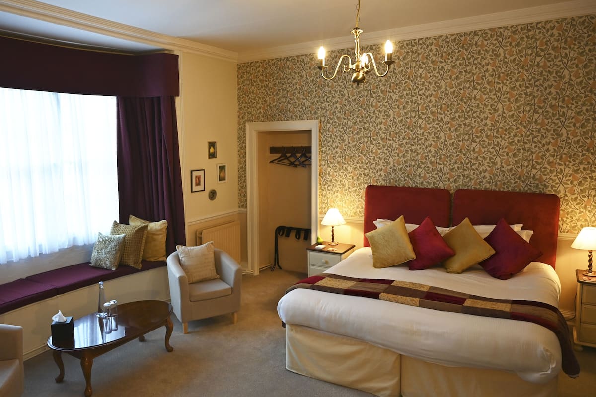 Ein Hotelzimmer mit einem großen Bett, einer Sitzecke inklusive Sessel und Tisch, einer Chandelüster und einem fenster mit Vorhängen.