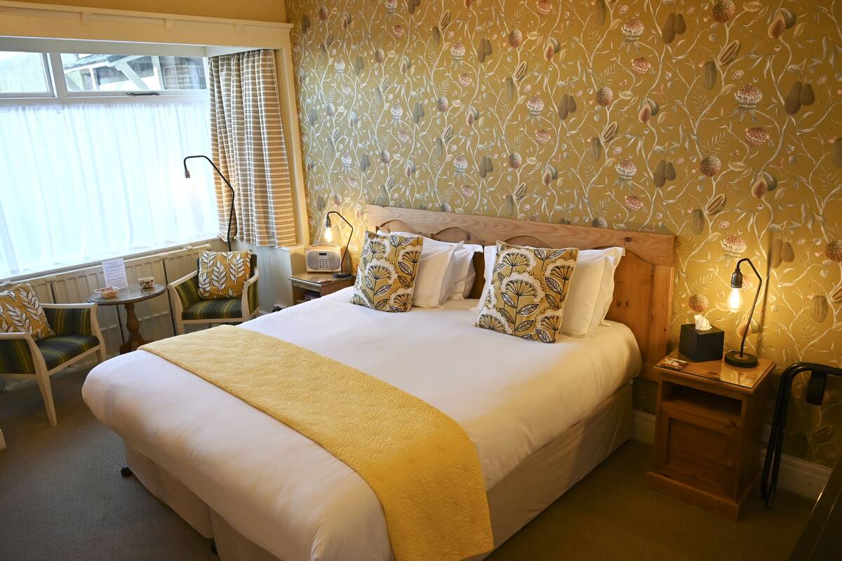 Ein Hotelzimmer mit einem großen Bett, einem Nachttisch mit Lampe, einem Sessel, einem kleinen Tischchen mit Radio und einem Fenster mit Vorhängen.