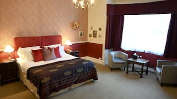 A hotel room with a large bed, two bedside tables with lamps, a small table with a chair, and a large window with curtains.
