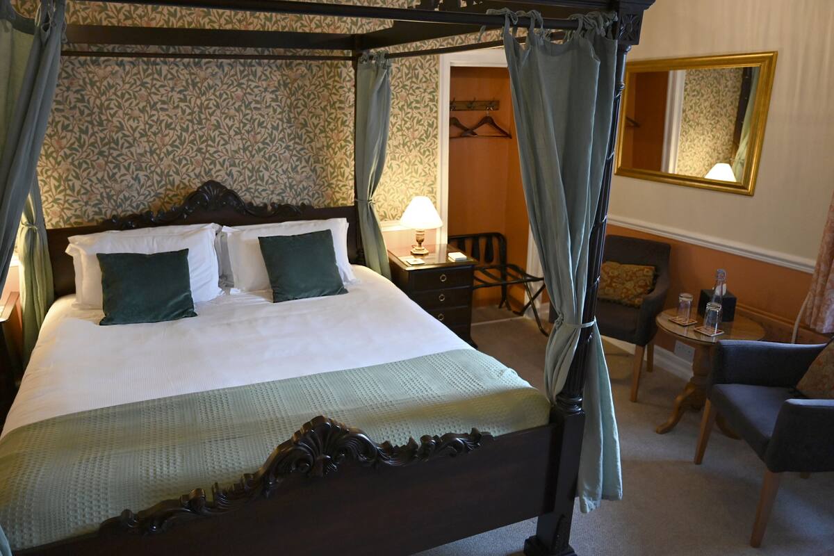 Ein Bett mit Himmelbett, ein Schreibtisch mit Lampe, ein Sessel und ein kleiner Tisch mit Gläsern.