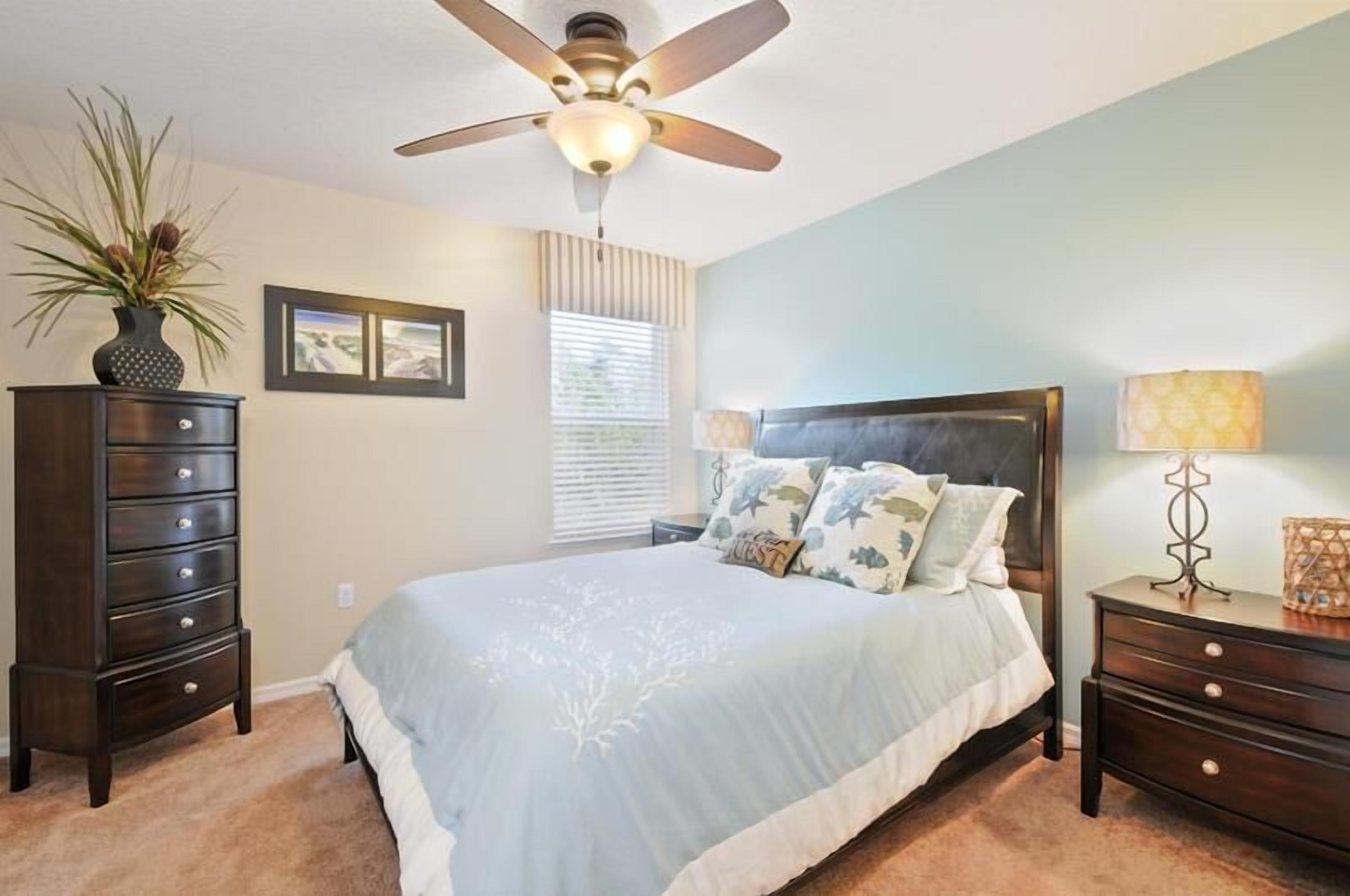 A bedroom with a bed, a ceiling fan, a dresser, a lamp, and a plant.