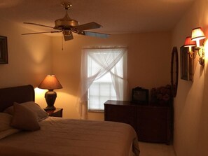 A bedroom with a bed, a nightstand with a lamp, a dresser, a ceiling fan, a window with curtains, and wall-mounted lamps.