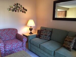 A living room with a patterned armchair, a green sofa with patterned cushions, a side table with a lamp, and a wall-mounted decorative piece. - Fantasy World Club Villas (Kissimmee)