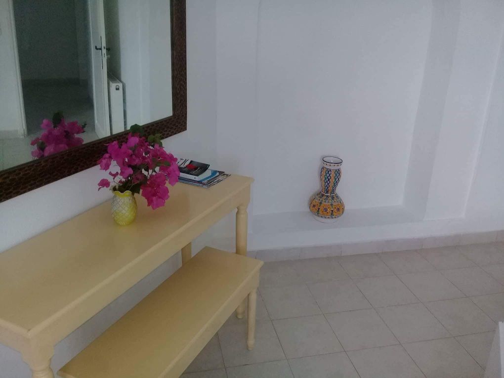 A room with a wooden bench, a mirror, a vase with flowers, and a decorative vase on the floor.
