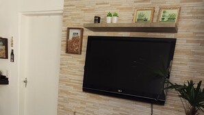 A living room with a flat-screen TV mounted on a textured wall, a small round table with a red top, a brown ottoman, and a potted plant. - Casa com piscina em Caraguatatuba (Caraguatatuba)