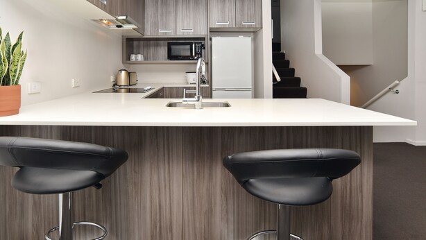 A modern kitchen with a large island, two black bar stools, a microwave, a refrigerator, and a staircase leading to another level. - Armagh Apartment 3 - Central Christchurch (Christchurch)