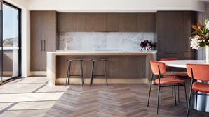 A modern kitchen with a herringbone floor pattern, wooden cabinets, a marble backsplash, and a central island with stools. - Carmel at Sorrento (Sorrento)