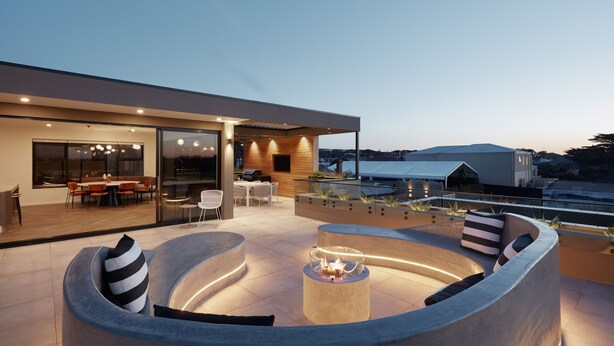 A modern outdoor living area with a circular seating arrangement, a fire pit, and a glass-enclosed dining space. - Carmel at Sorrento (Sorrento)