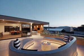 A modern outdoor living area with a circular seating arrangement, a fire pit, and a glass-enclosed dining space. - Carmel at Sorrento (Sorrento)