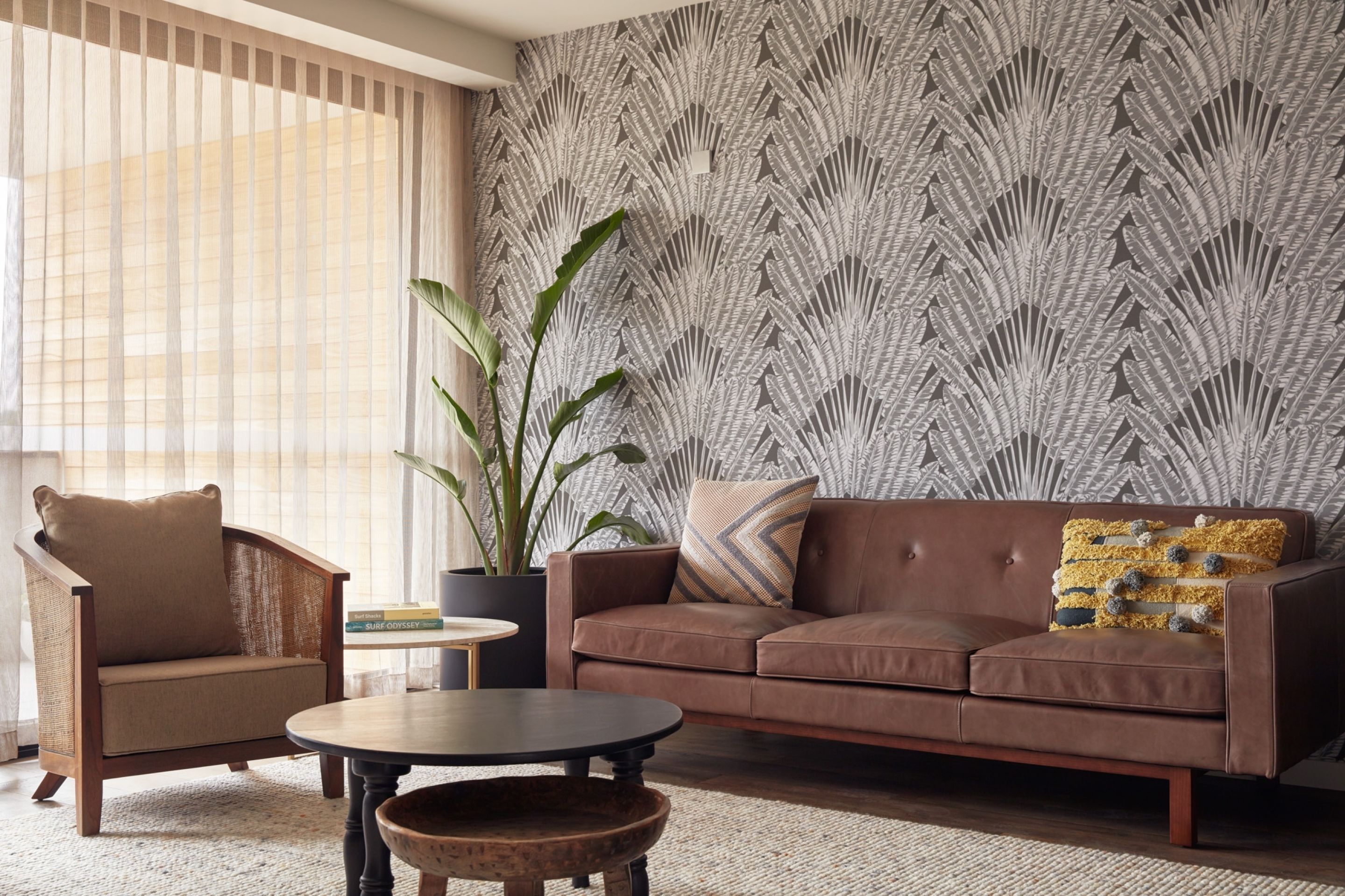 A living room with a brown sofa, a round coffee table, and a wicker armchair. There is a large potted plant and a patterned wallpaper on the wall.