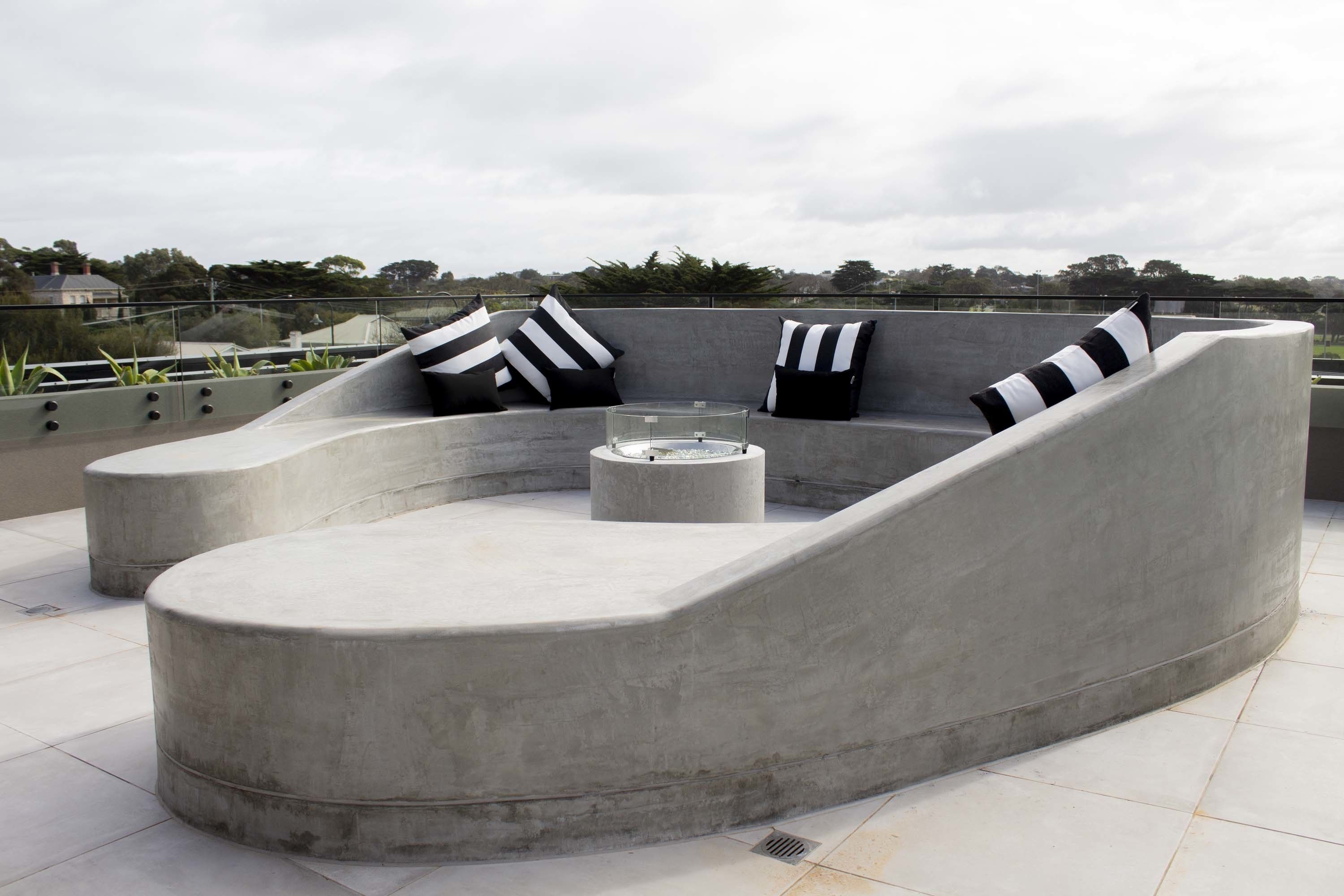 A modern outdoor seating area with a curved concrete bench and a glass-top table, featuring black and white striped cushions.
