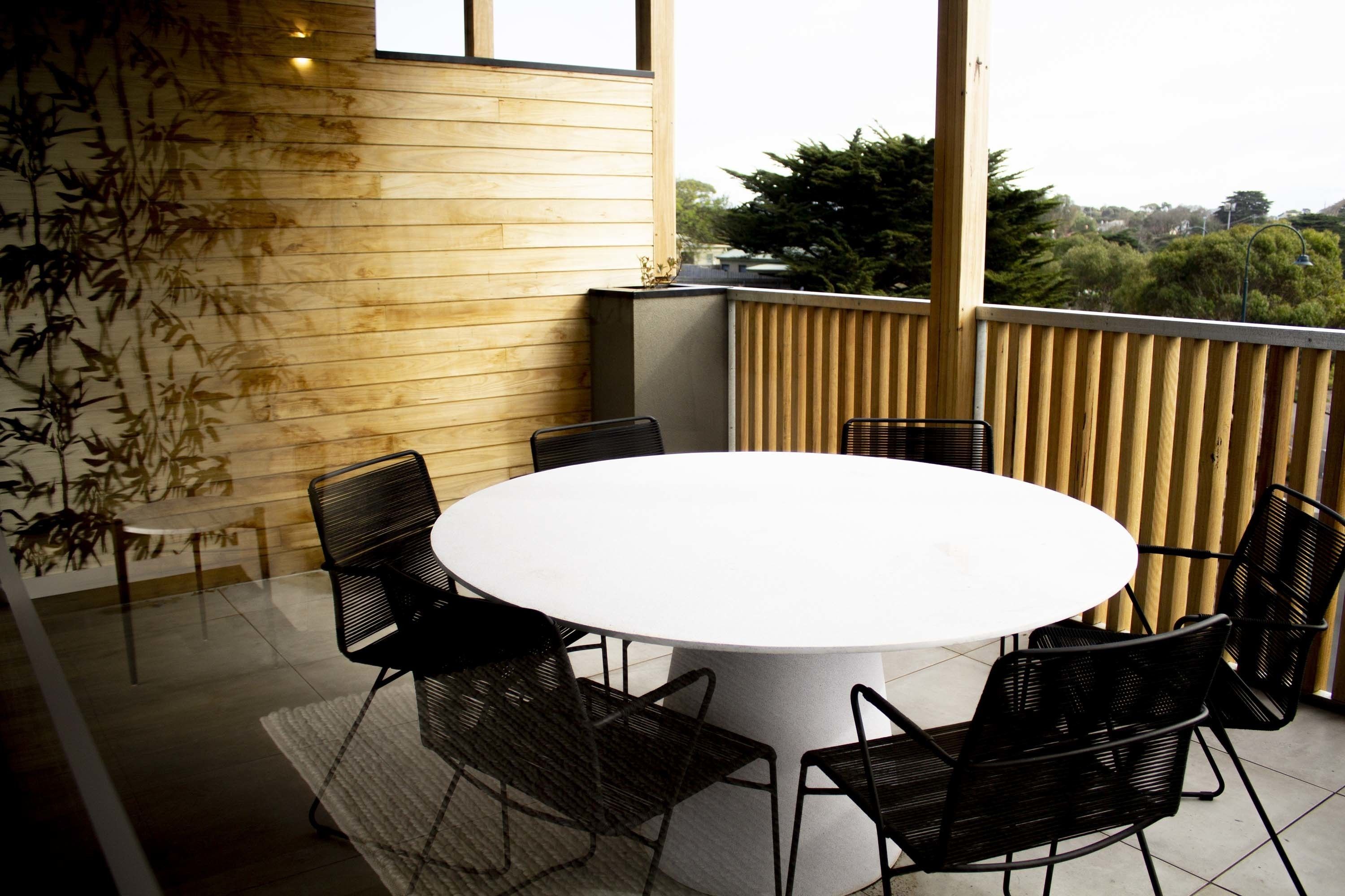 A round table with six chairs in an outdoor setting with a wooden wall and a view of trees.
