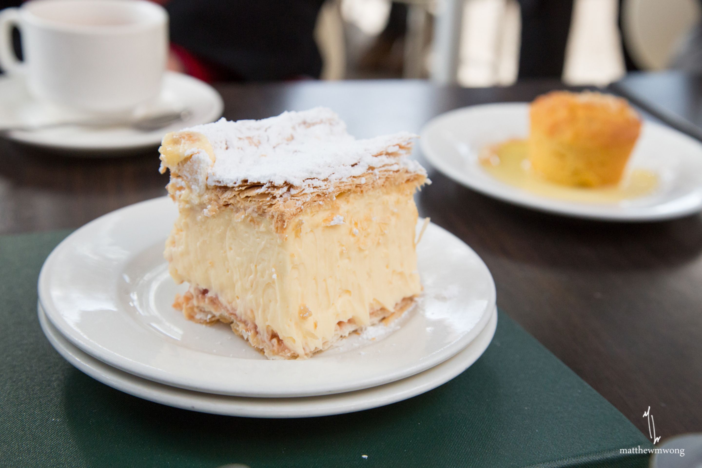 A slice of cake on a plate with powdered sugar on top, a muffin with syrup, and a cup of coffee on a saucer.