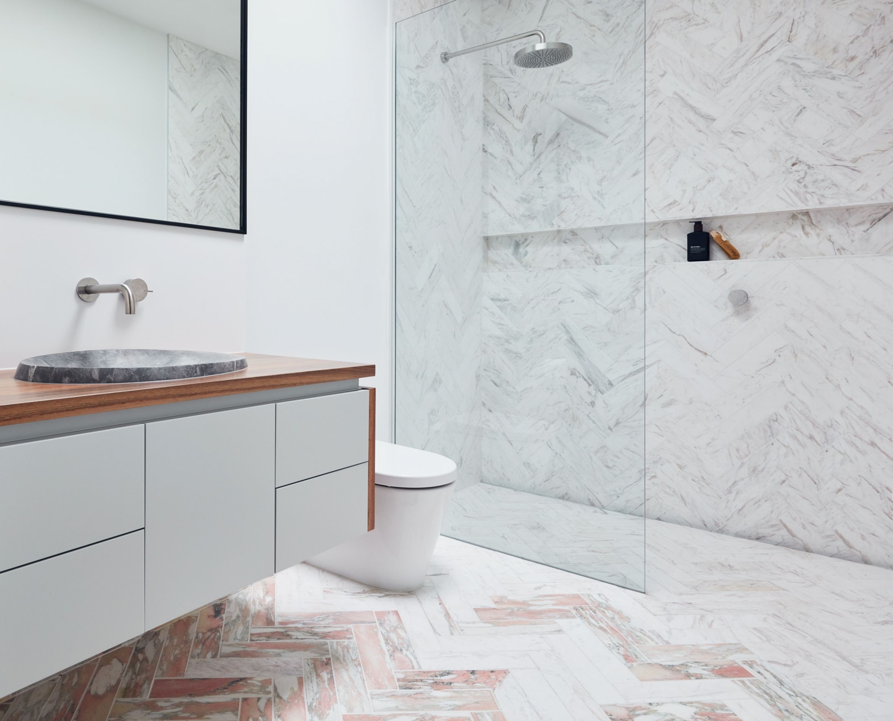 A modern bathroom with a marble shower wall, a wooden vanity, and a herringbone-patterned floor.