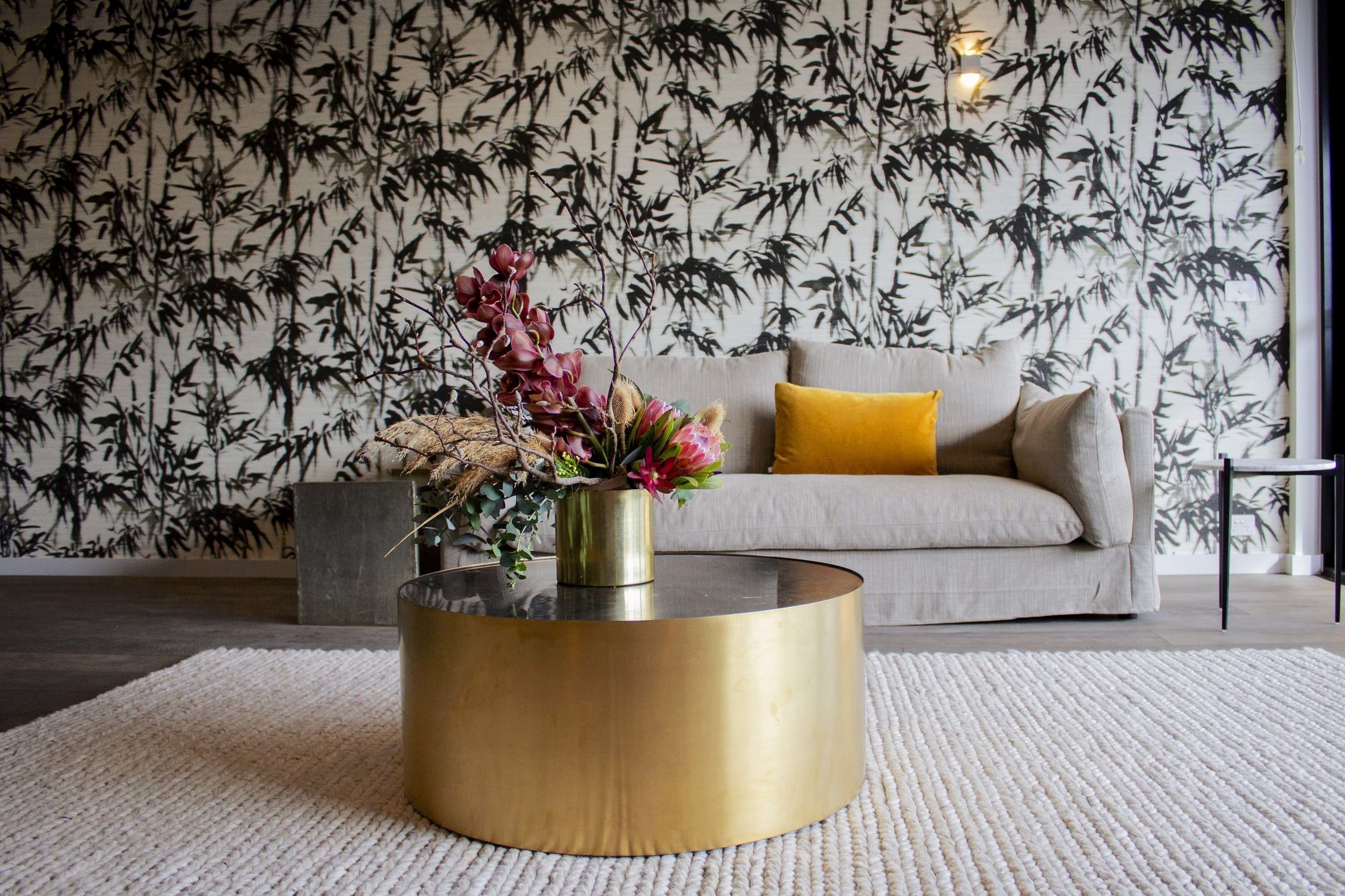 A living room with a round gold coffee table, a beige sofa with a mustard yellow pillow, and a wall with a bamboo pattern.
