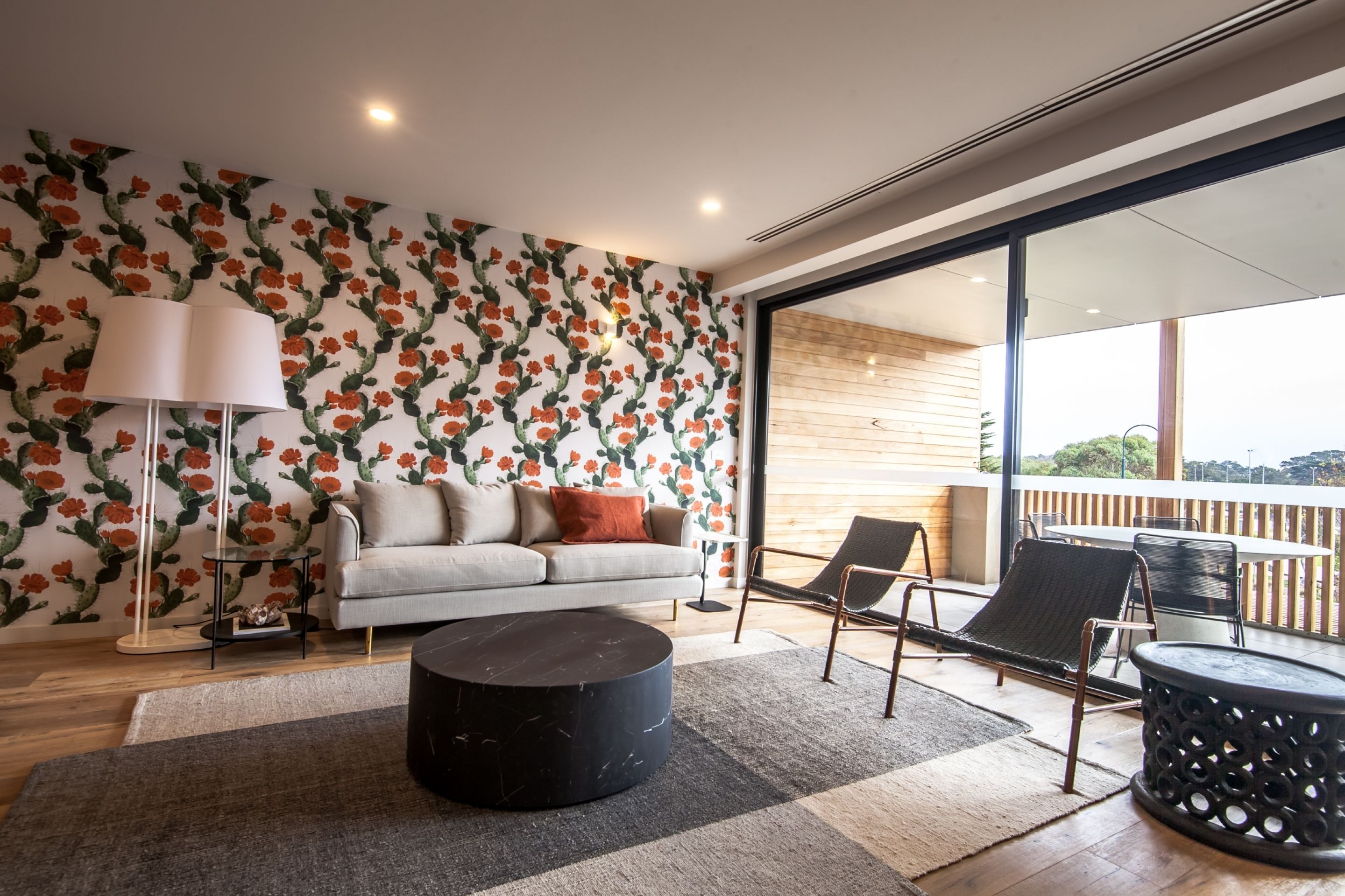 A modern living room with a sofa, a coffee table, and a floor lamp. There is a large window with a view of the outdoors.