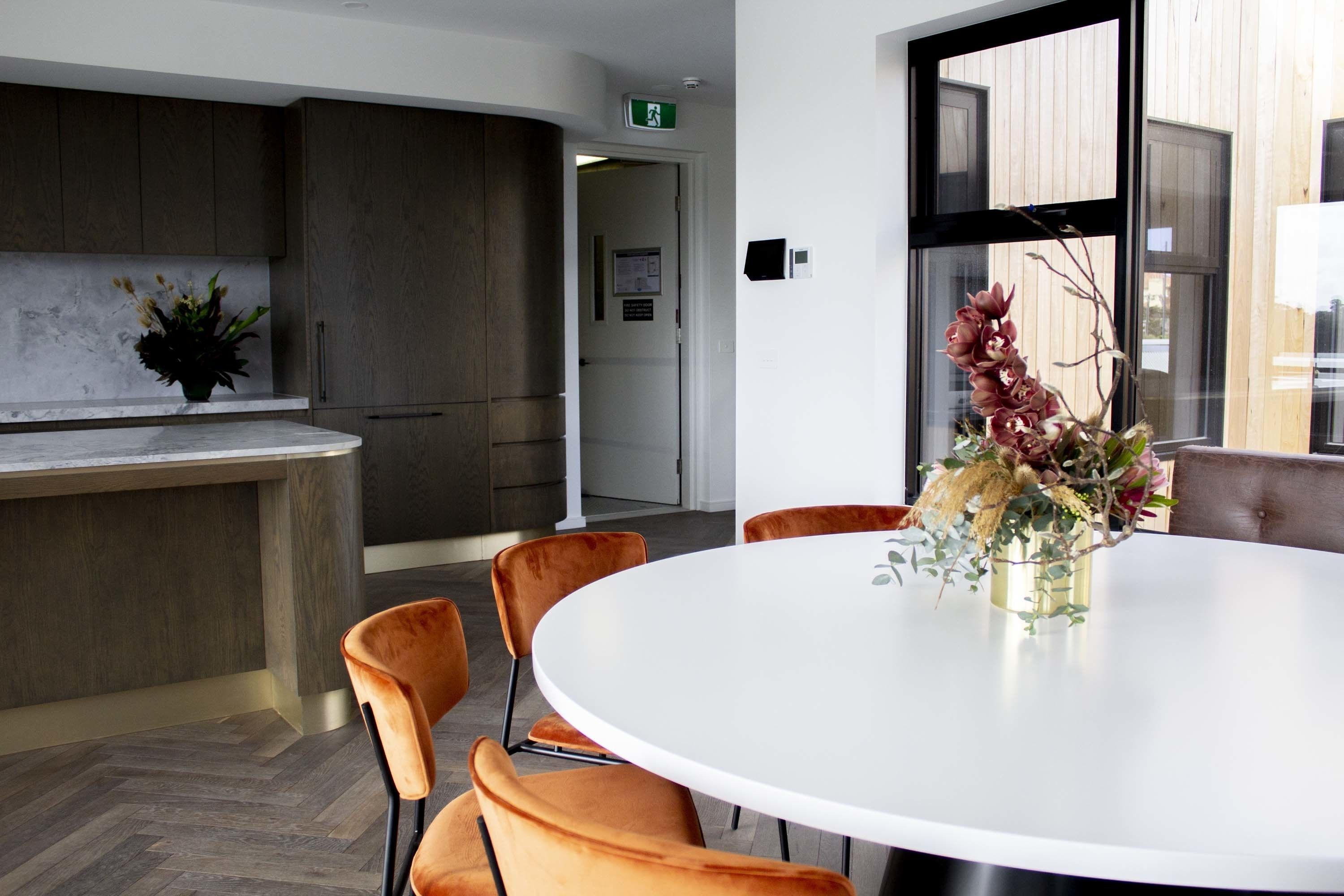 A modern kitchen with dark wood cabinets, a marble countertop, and a round table with orange chairs.