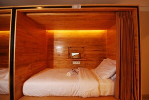 A wooden cabin-style room with a single bed, a small shelf with a television, and a curtain. - Dbeds Hostel By Soscomma (Kuta)