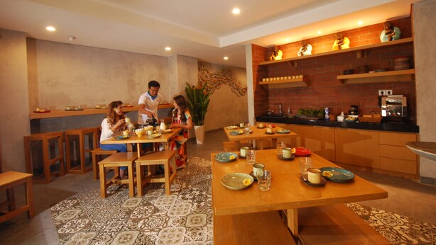 A dining area with multiple wooden tables and chairs, a kitchen area with brick walls and shelves, and a patterned floor. - Dbeds Hostel By Soscomma (Kuta)