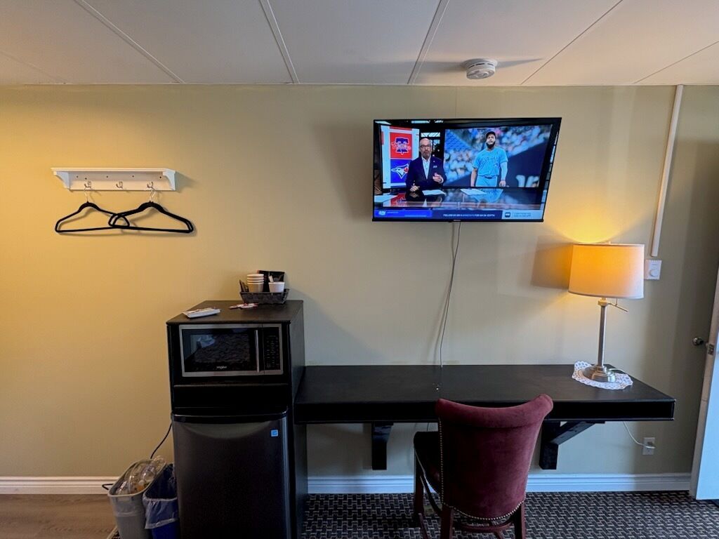 A hotel room with a desk, a television mounted on the wall, a chair, a trash bin, and a coat rack.