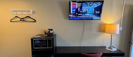 A hotel room with a desk, a television mounted on the wall, a chair, a trash bin, and a coat rack.