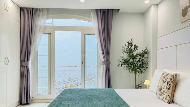 Ein Schlafzimmer mit Bett, Vorhängen, einer Pflanze und Blick durch ein Fenster.