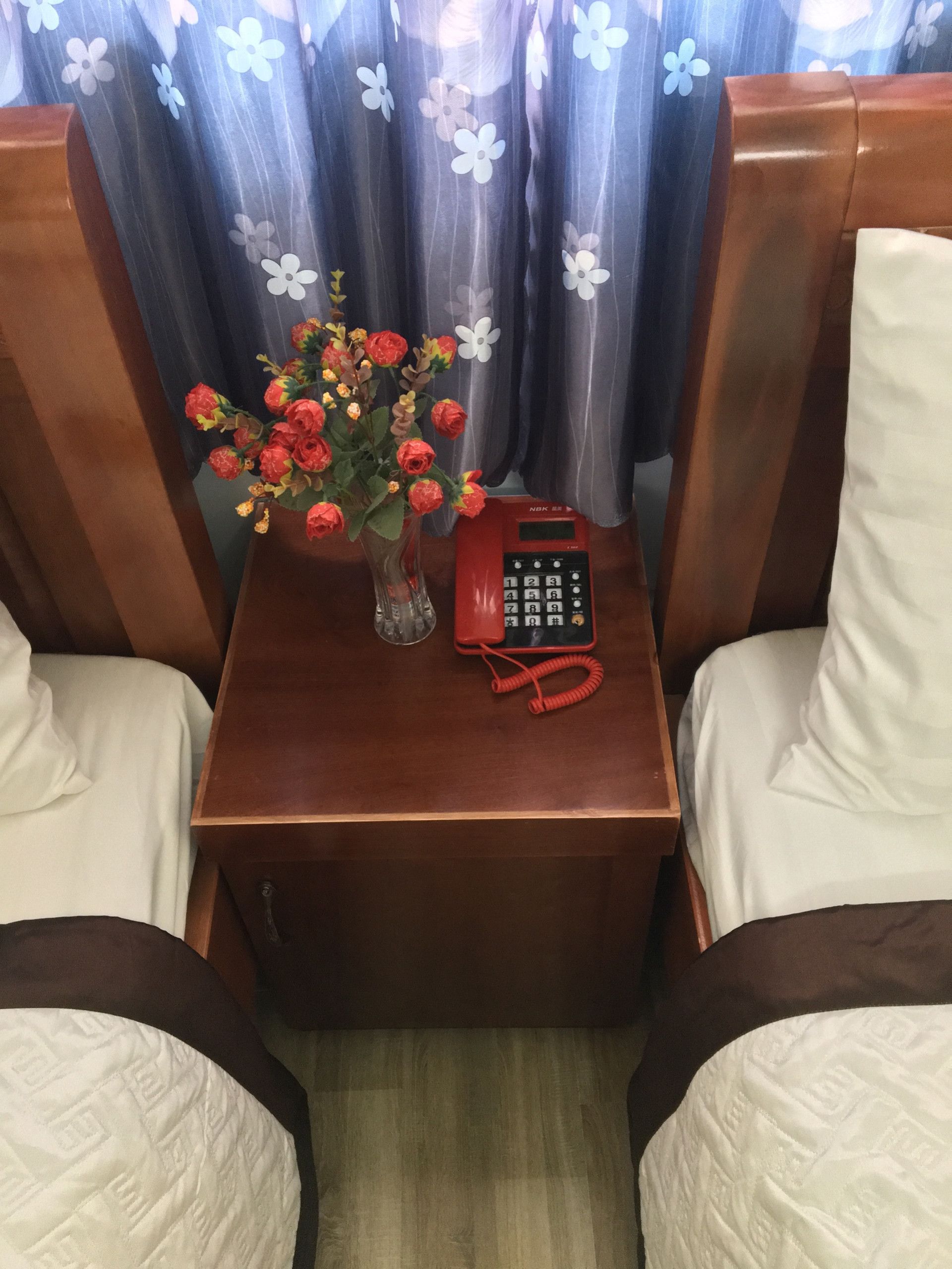 Een houten nachtkastje met een vaas rode bloemen, een rode telefoon en een bruine stoel met een witte kussentje.