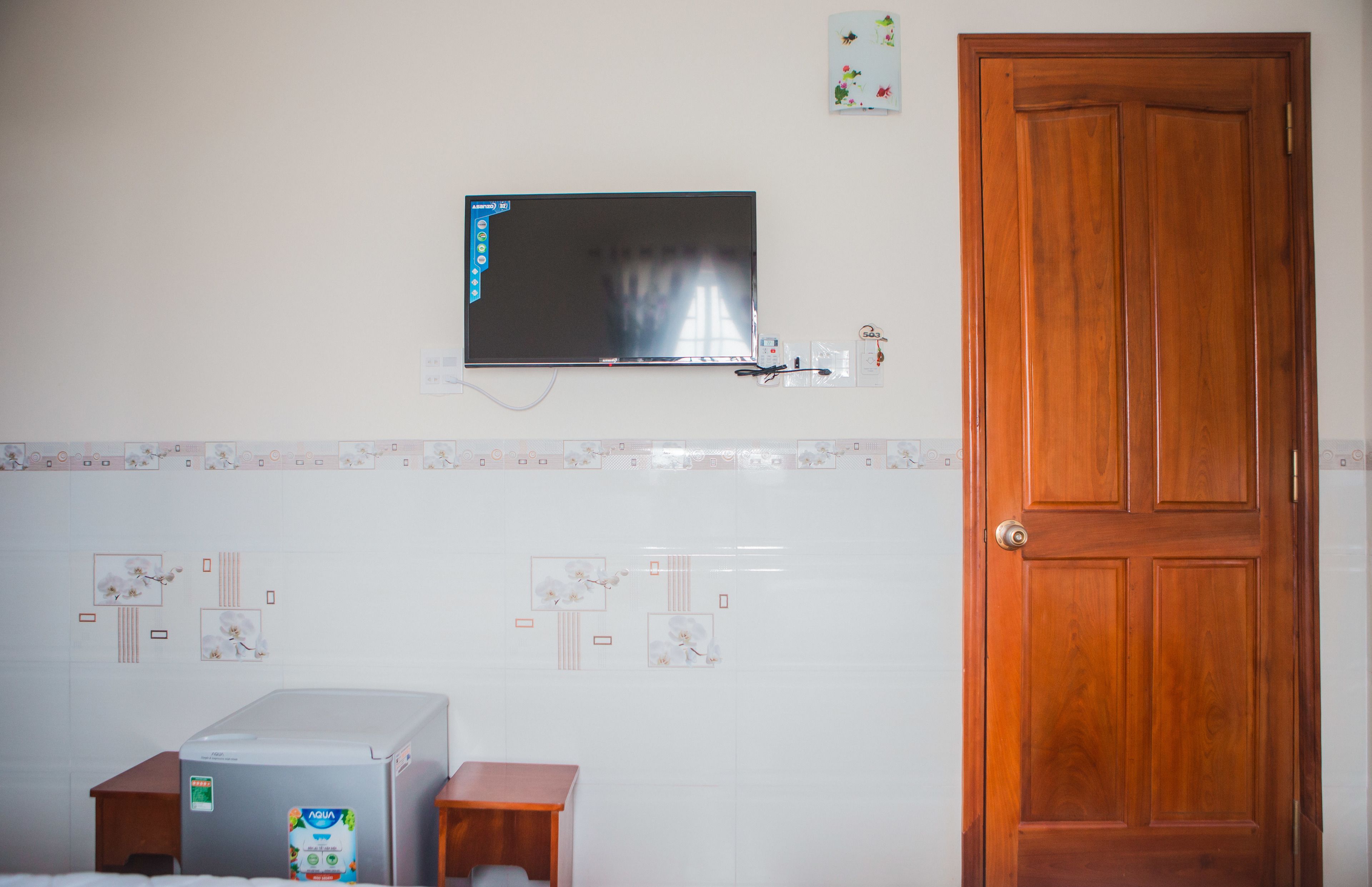 A room with a wooden door, a mounted television, a refrigerator, and a tiled backsplash with floral designs.