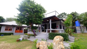 A two-story house with a covered porch, a staircase leading to the upper floor, and a large tree in the front yard. - V Garden (Koh Samui)