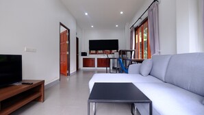 A modern living room with a sofa, a TV on a wooden stand, a small table, and a kitchen area in the background. - V Garden (Koh Samui)