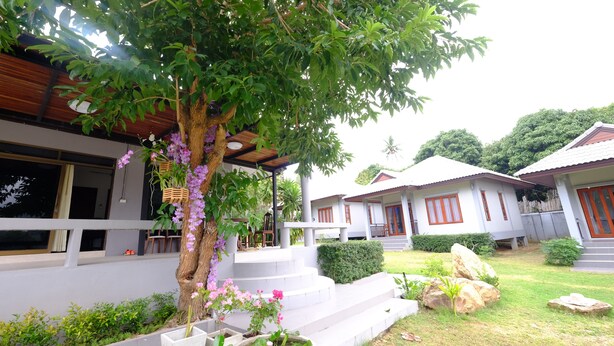 A bungalow with a wooden porch, steps leading to the entrance, and a garden with a tree and flowers. - V Garden (Koh Samui)
