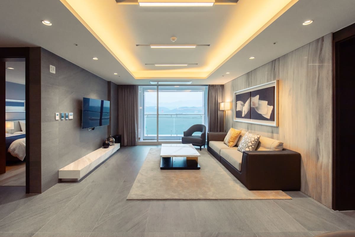 Ein modernes Hotelzimmer mit einem großen Bett, einer Couch, einem Couchtisch und einem Fernseher. Das Zimmer verfügt über ein großes Fenster mit Blick auf das Meer und die Berge.