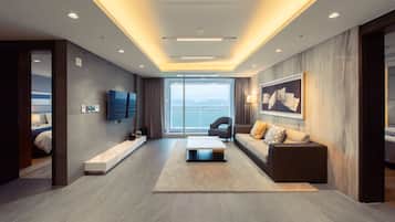 Habitación de hotel moderna con una cama grande, un sofá, una mesita de café y un televisor. La habitación tiene un ventanal con vistas al mar y a las montañas.