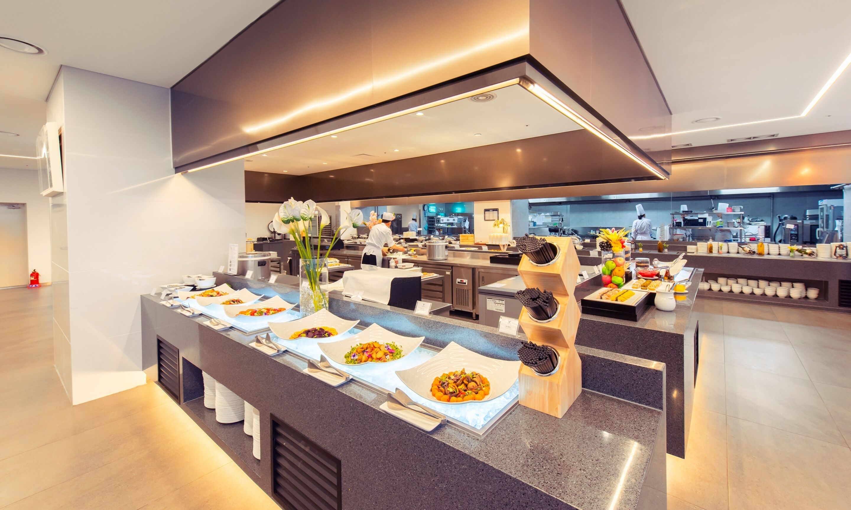 Un moderno bufé de desayuno con una variedad de platos, una zona de servicio central y una cocina al fondo.