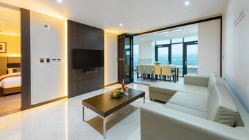 A modern hotel room with a bed, a TV, a dining area with a table and chairs, and a balcony with a view of the sea.