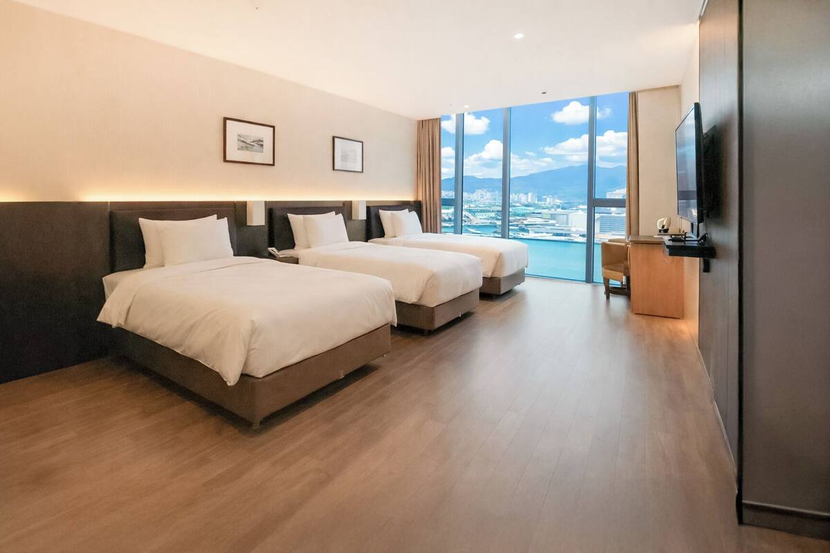 Ein Hotelzimmer mit zwei Betten, einem Schreibtisch und einem großen Fenster mit Blick auf die Stadt.