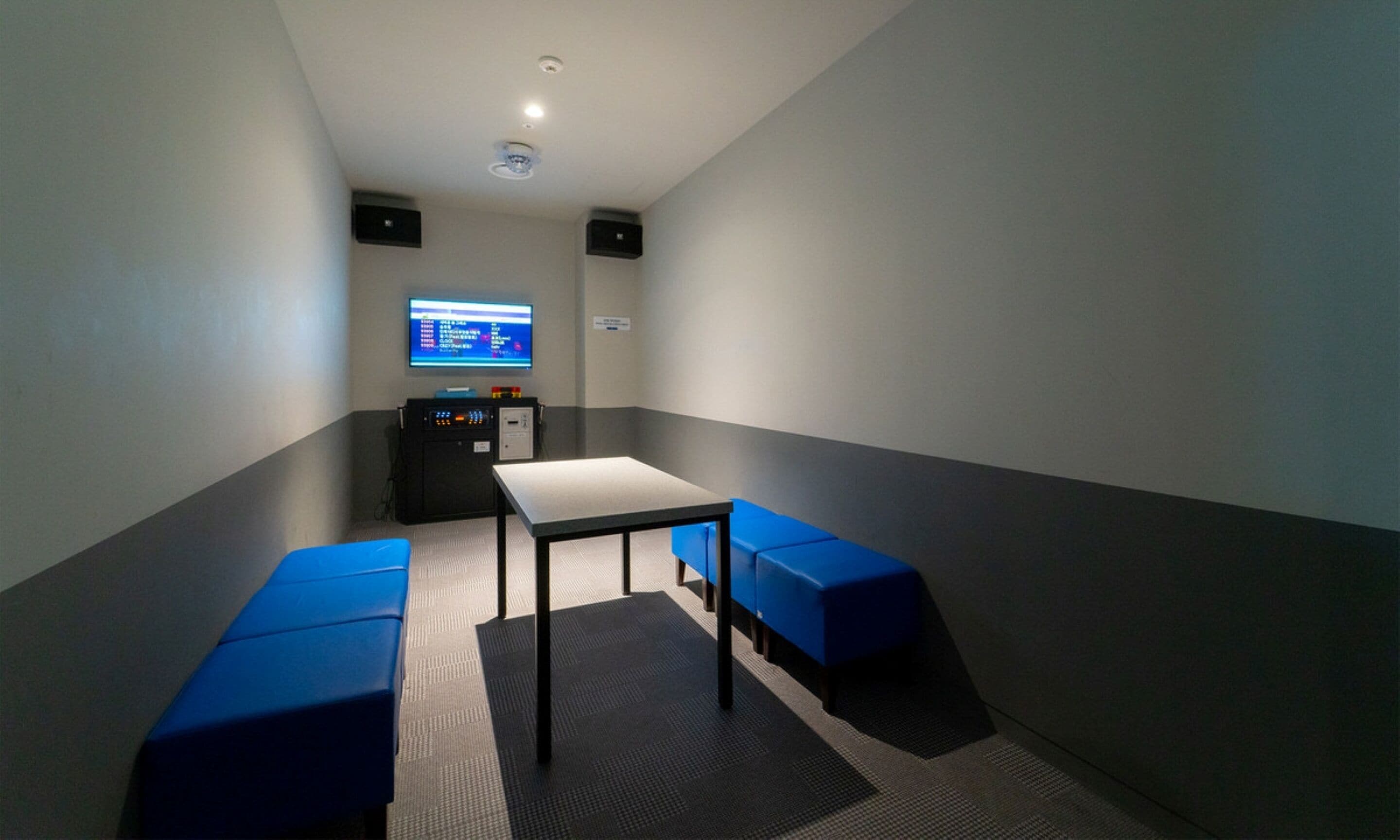 Pequeña habitación tenuemente iluminada, con una mesa, dos bancos azules, un televisor montado en la pared y un mueble pequeño con un panel de control.