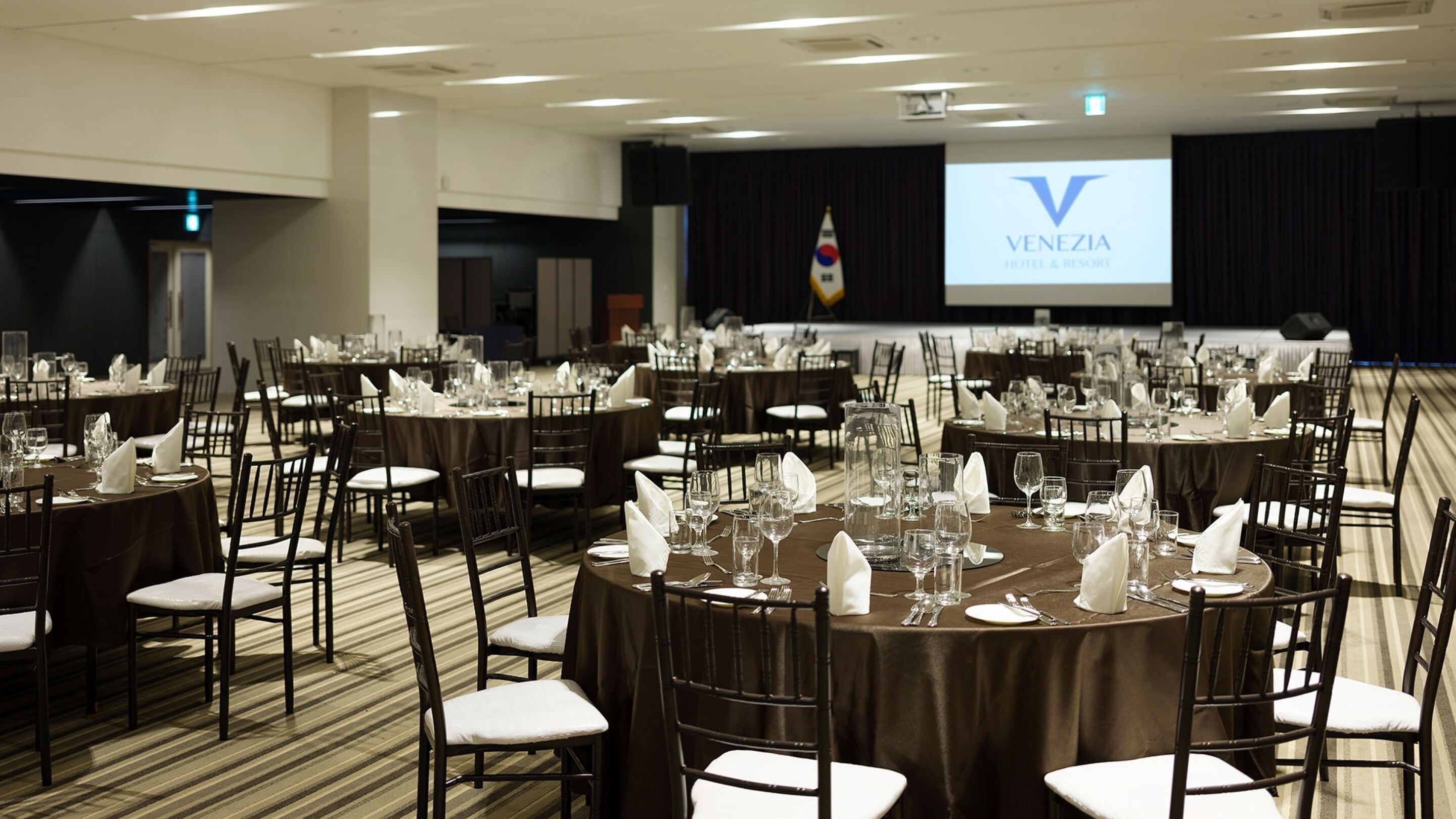 A conference room with round tables set for a formal event, a projector screen displaying the Venezia Hotel & Resort logo, and a flag in the background.