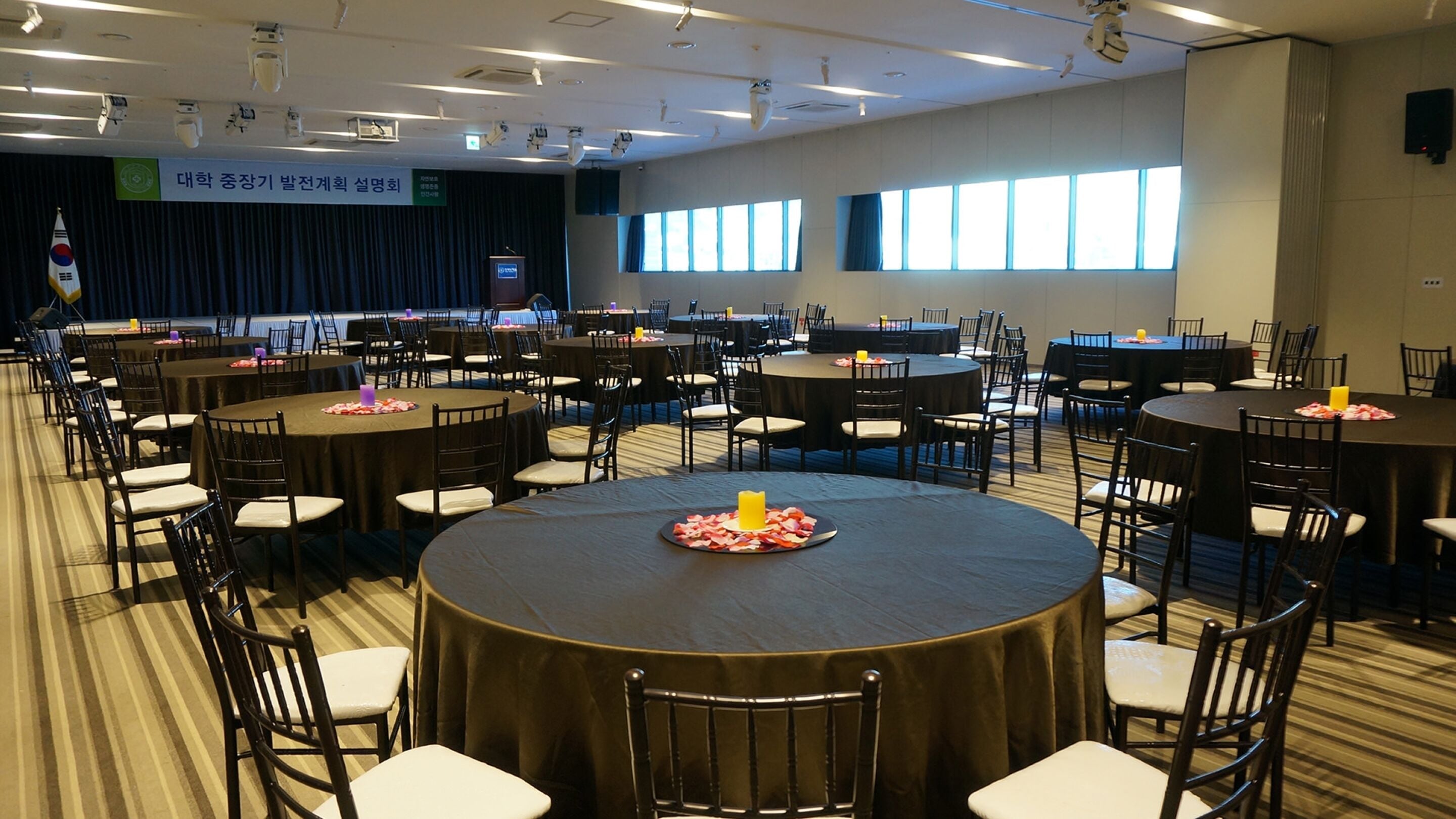 Une salle de conférence dotée de plusieurs tables rondes et de chaises, de grandes fenêtres et d’une scène sur laquelle flotte un drapeau.